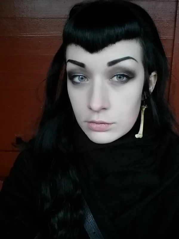 Portrait de profil avec cheveux noirs, maquillage sombre, col roulé noir, boucle d'oreille os, fond bois