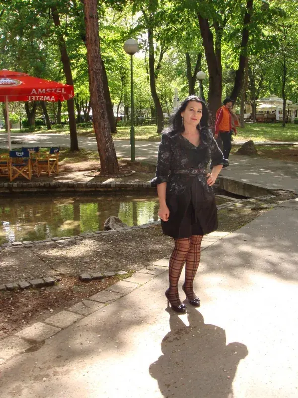 Personne en robe noire à motifs, collants à mailles et talons, debout sur un chemin de parc ombragé près d'un étang, sous un parasol rouge.