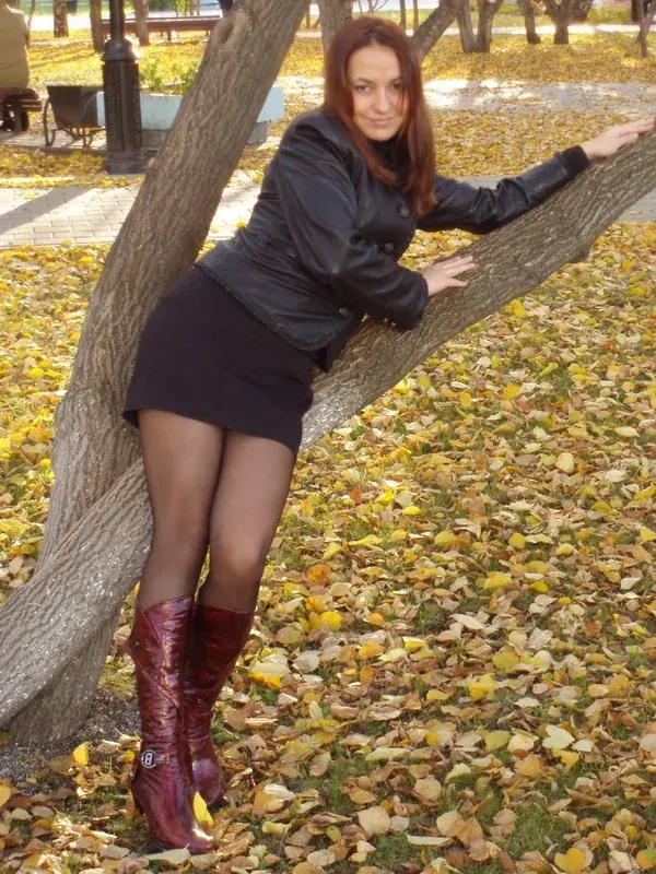 Personne appuyée sur un arbre en automne, feuilles mortes au sol, veste noire, jupe courte, bottes bordeaux vernies