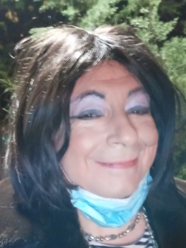 Portrait de profil, cheveux foncés, maquillage yeux, masque bleu sous menton, veste sombre, haut rayé, collier métallique, fond végétal flou