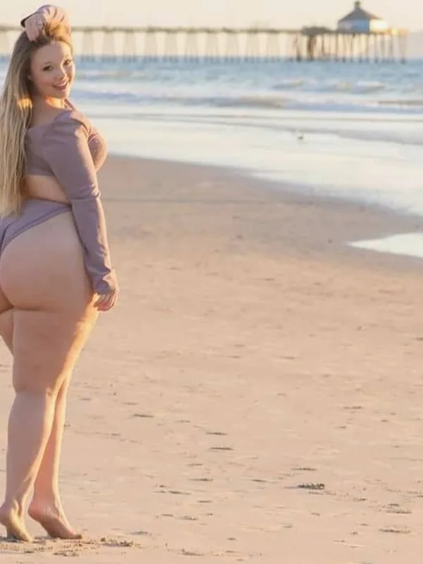 Personne en tenue mauve sur une plage, dos tourné, regardant l'objectif, avec océan et ponton en arrière-plan.