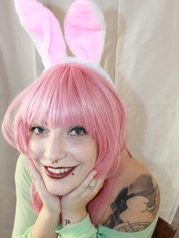 Portrait de personne avec perruque rose, oreilles de lapin, pull vert, tatouages, mains sur le visage, sourire, fond clair
