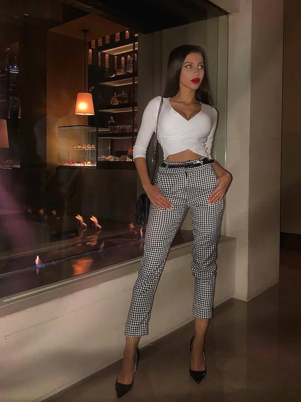 Personne en tenue élégante, haut blanc, pantalon à carreaux, escarpins noirs, posant près d'une vitrine éclairée.