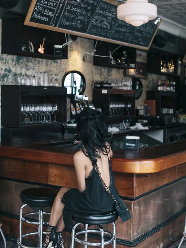 Personne de dos assise sur un tabouret au comptoir d'un bar, robe noire, cheveux longs, lumière chaude, décor rustique