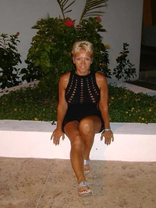 Femme aux cheveux blonds courts assise sur un rebord blanc, vêtue d'une robe noire à découpes, devant des plantes vertes.
